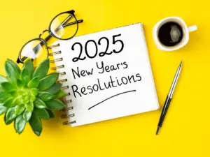 A notepad with '2025 New Year's Resolutions' written on it, accompanied by a pen, glasses, a plant, and a cup of coffee—symbolizing planning and reflection for psychotherapists exploring the best therapy training programs in 2025