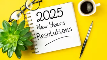 A notepad with '2025 New Year's Resolutions' written on it, accompanied by a pen, glasses, a plant, and a cup of coffee—symbolizing planning and reflection for psychotherapists exploring the best therapy training programs in 2025